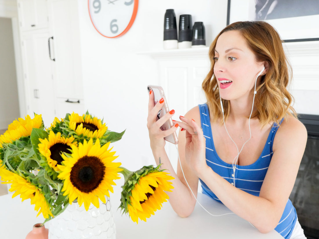 Eva Amurri Martino of blog REZTT sitting at her kitchen table next to a vase of sunflowers, wearing a striped blue and white maternity tank and listening to an audiobook on Audible