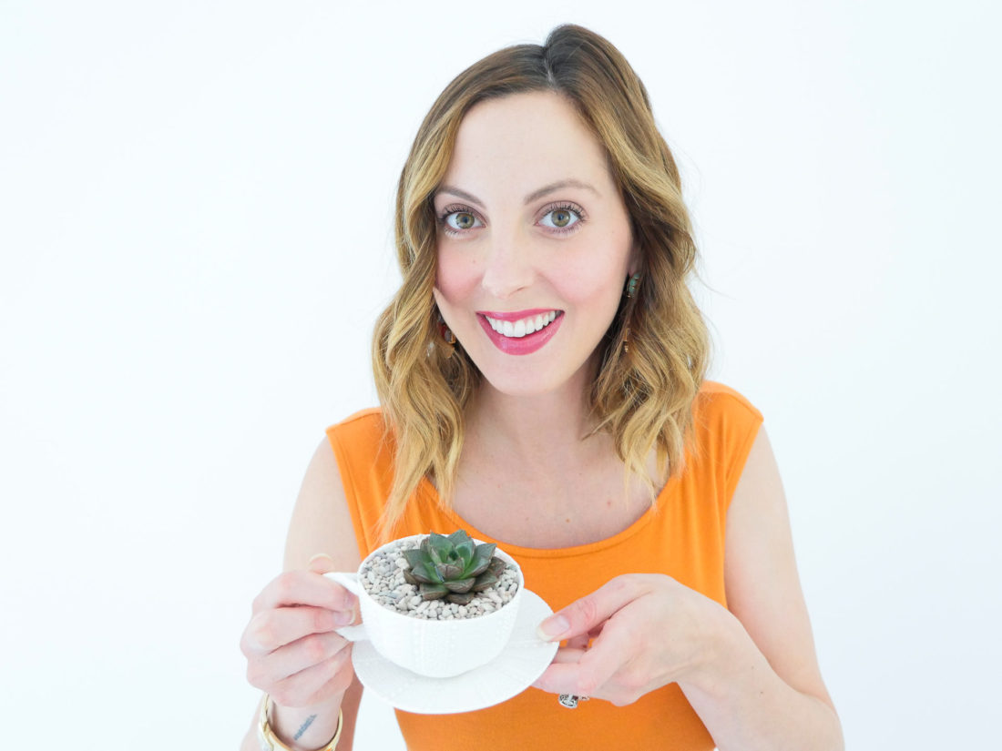 Eva Amurri Martino from the REZTT blog displaying her DIY Teacup Planters with Mini Succulents and small pebbles, wearing a bodycon orange maternity dress and gold bracelet watch