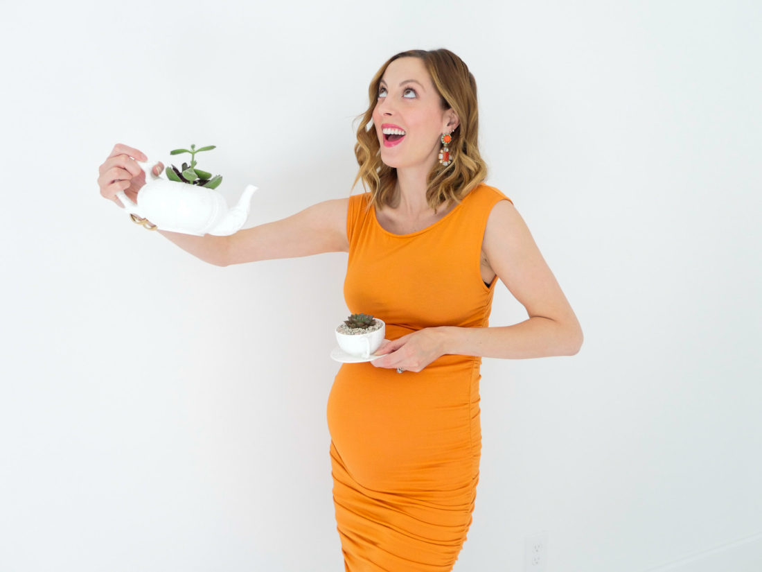 Eva Amurri Martino from the REZTT blog displaying her DIY Teacup Planters with Mini Succulents and small pebbles, wearing a bodycon orange maternity dress, at thirty three weeks pregnant, colorful chandelier earrings, and gold bracelet watch