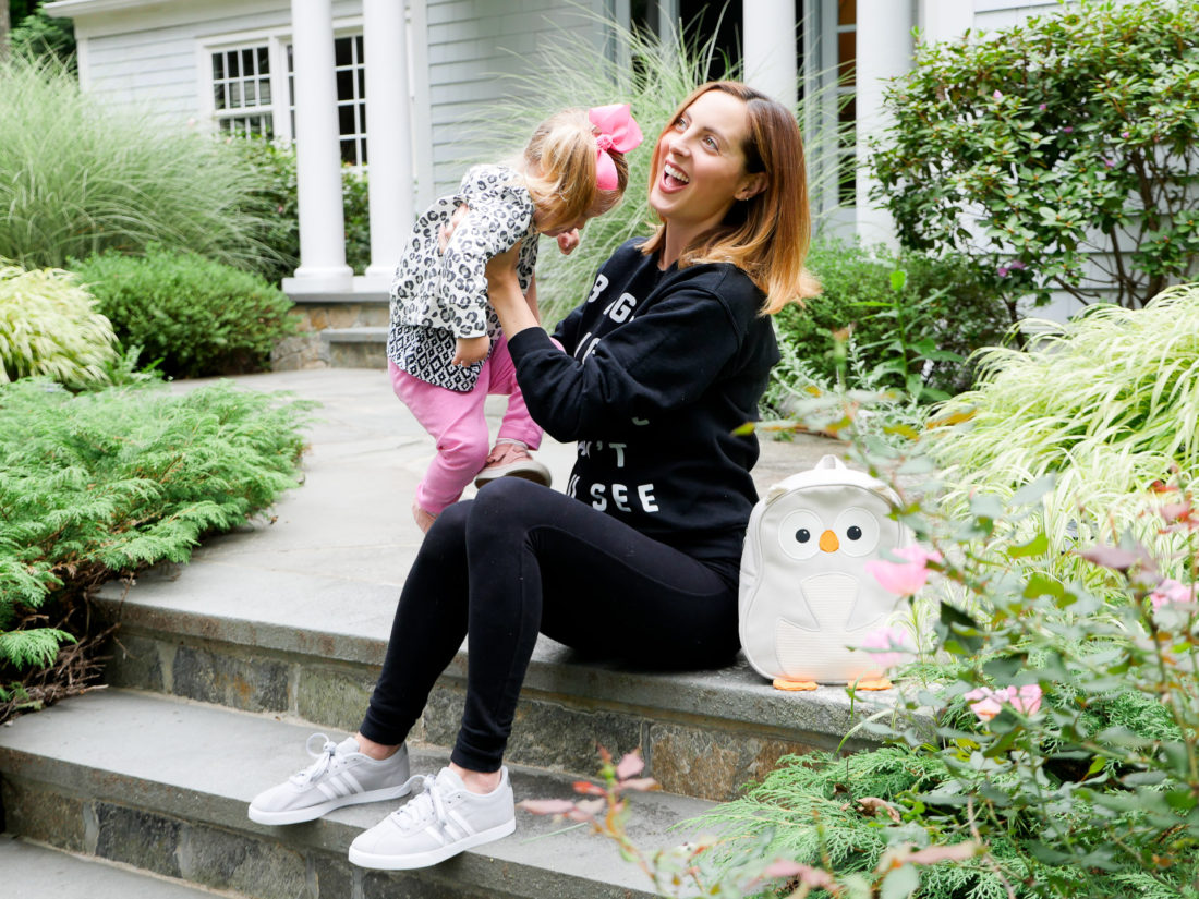 Eva Amurri Martino of lifestyle blog REZTT sitting on the steps of her connecticut home wearing black maternity leggings, a blac sweatshirt, and light grey adidas sneakers, lifting her daughter Marlowe on to her lap