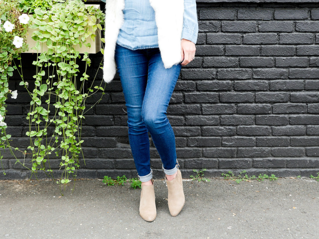 Eva Amurri Martino of REZTT blog wearing maternity jeans, a chambray maternity top, a white fur vest, and tan colored fall boots standing against a dark grey wall