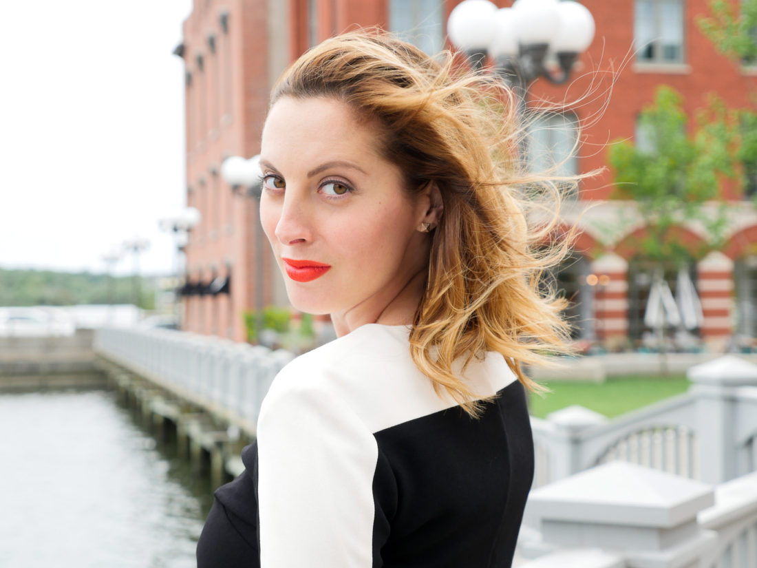 Eva Amurri Martino of lifestyle blog REZTT wearing red lipstick and a cream and black dress standing at the waterfront in connecticut and looking back over her shoulder at the camera