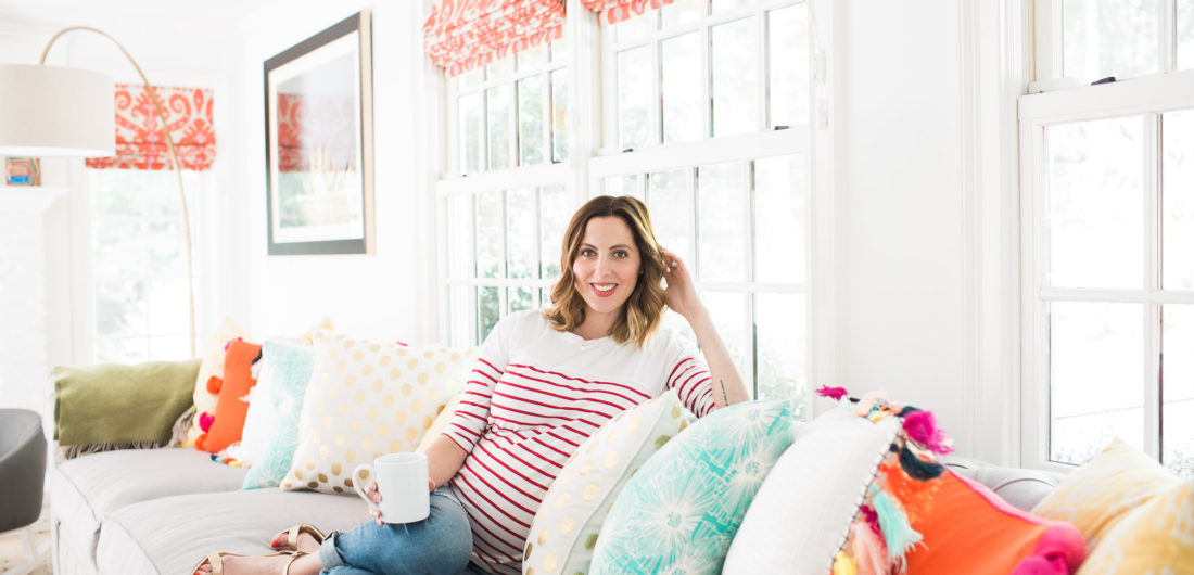 Eva Amurri Martin of lifestyle blog REZTT relaxes at home in her family room on a grey couch with multicolored and multipatterned pillows surrounding her, holding a cup of coffee