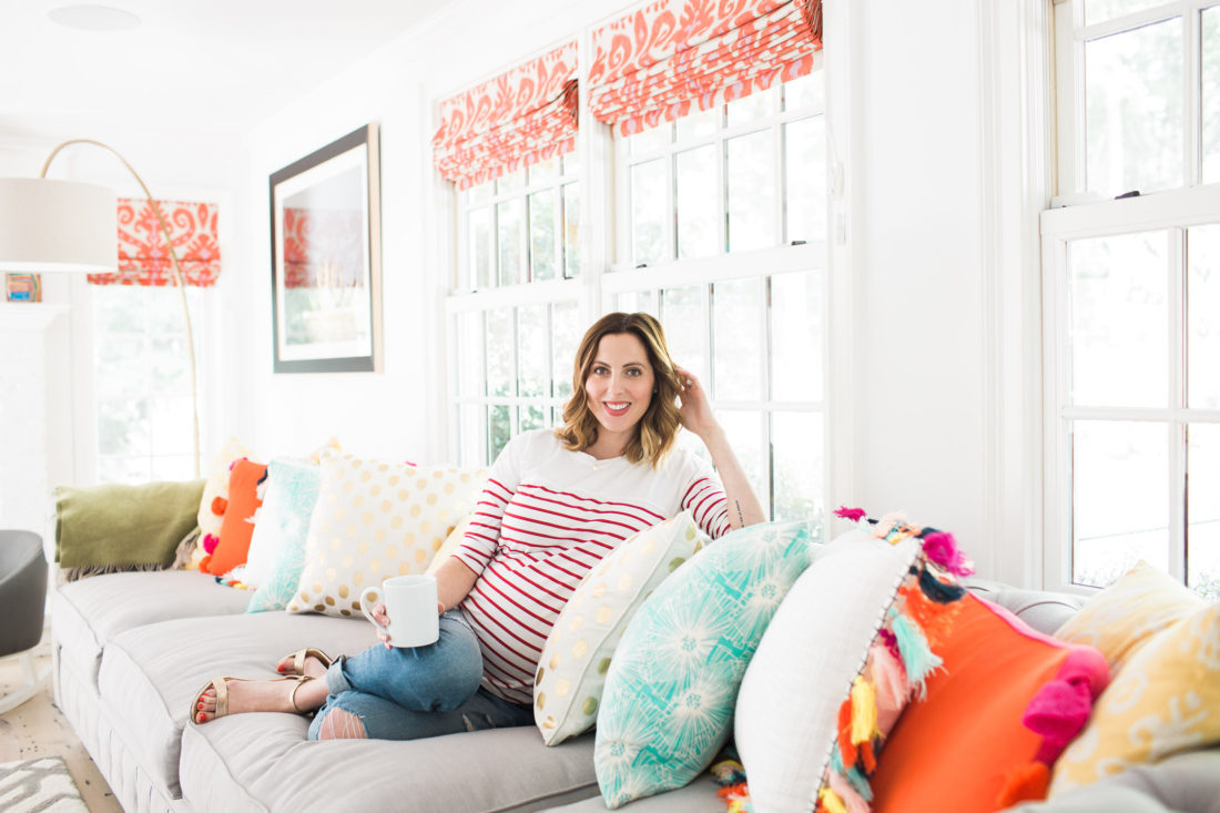 Eva Amurri Martin of lifestyle blog REZTT relaxes at home in her family room on a grey couch with multicolored and multipatterned pillows surrounding her, holding a cup of coffee