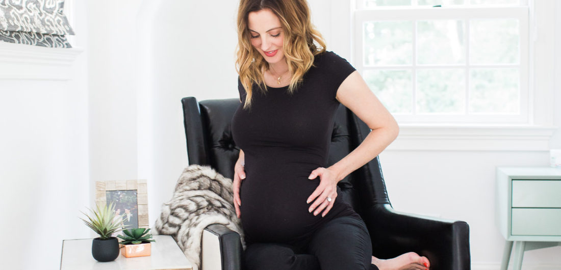 Eva Amurri Martino of lifestyle blog REZTT cradling her 37 week bump while sitting in a black leather chair in her master bedroom next to a brass table and two succulents
