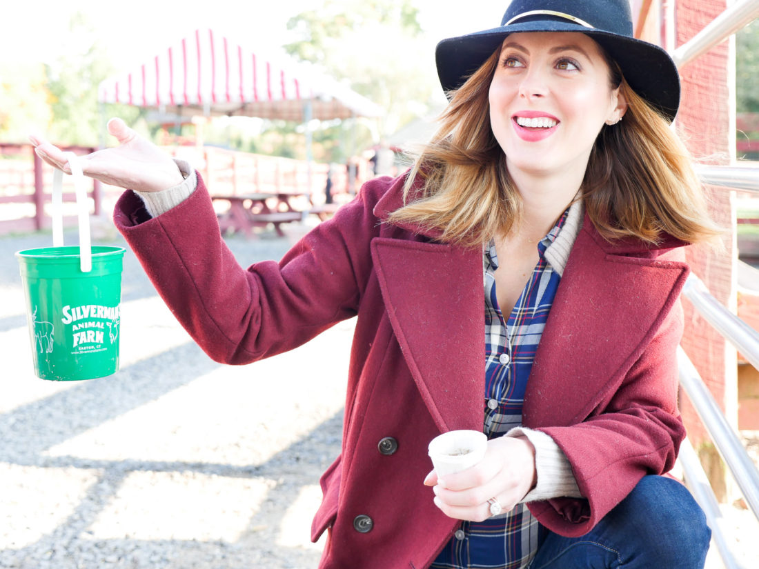 Eva Amurri Martino of lifestyle and motherhood blog REZTT, wearing a plaid shirt and oxblood wool peacoat, holding a feed bucket at Silverman's farm in connecticut