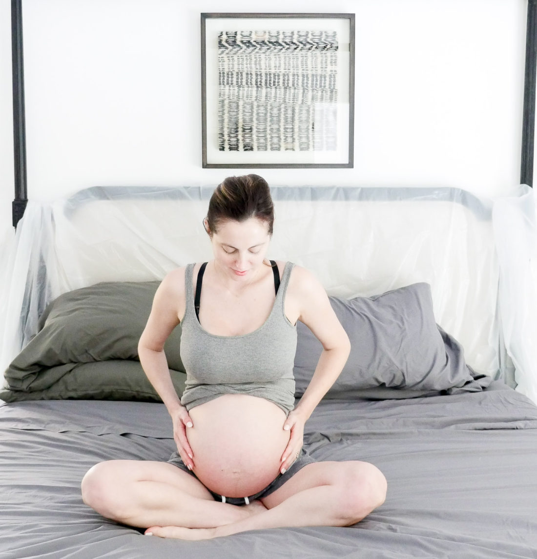 Eva Amurri Martino of lifestyle and motherhood blog REZTT in the early stages of labor at her home in connecticut, sitting on her bed in a grey tank top and grey shorts cradling her baby bump