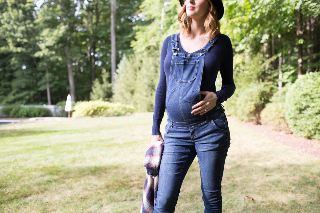 Eva Amurri Martino of lifestyle blog REZTT standing in her yard in maternity overalls, a navy felt hat, and a plaid poncho at her home in connecticut
