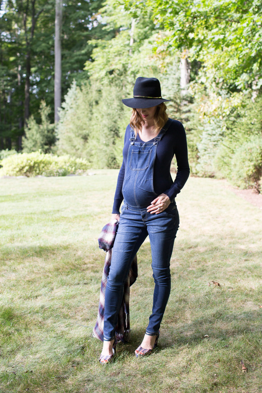 Eva Amurri Martino of lifestyle blog REZTT standing in her yard in maternity overalls, a navy felt hat, and a plaid poncho at her home in connecticut