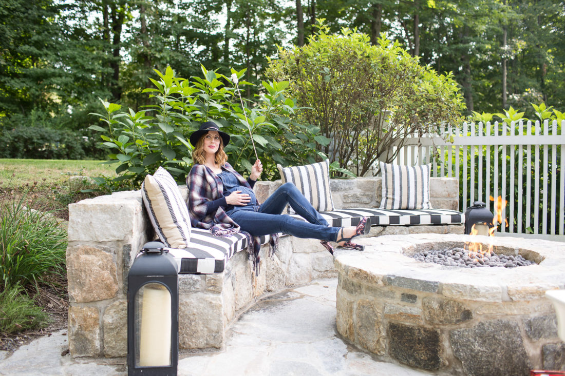 Eva Amurri Martino of lifestye and motherhood blog REZTT at her firepit at her connecticut home, wearing maternity overalls, a navy felt hat, and a plaid poncho roasting marshmallows over the fire