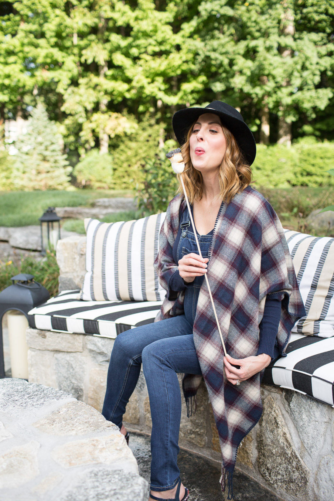 Eva Amurri Martino of lifestye and motherhood blog REZTT at her firepit at her connecticut home, wearing maternity overalls, a navy felt hat, and a plaid poncho roasting marshmallows over the fire