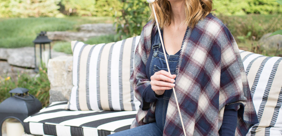 Eva Amurri Martino of lifestye and motherhood blog REZTT at her firepit at her connecticut home, wearing maternity overalls, a navy felt hat, and a plaid poncho roasting marshmallows over the fire