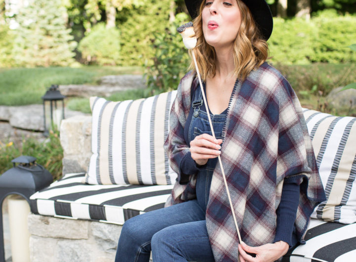 Eva Amurri Martino of lifestye and motherhood blog REZTT at her firepit at her connecticut home, wearing maternity overalls, a navy felt hat, and a plaid poncho roasting marshmallows over the fire