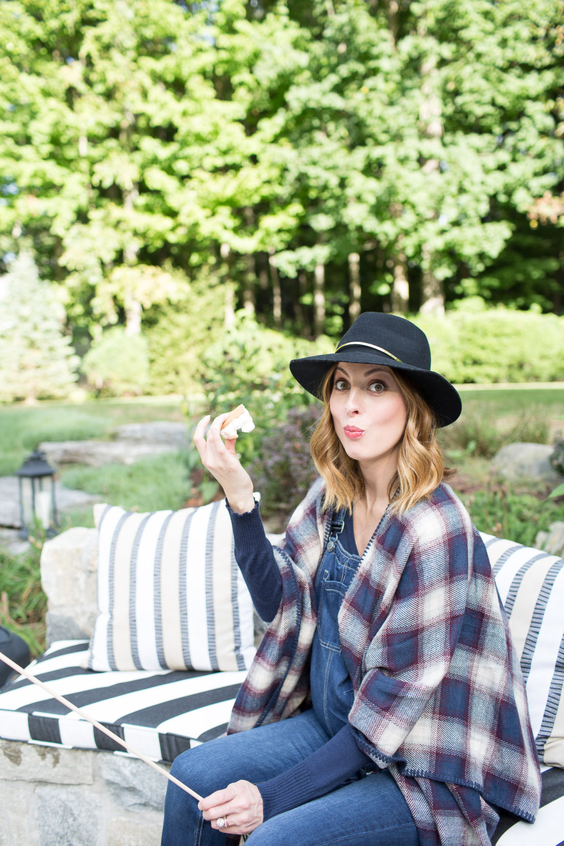 Eva Amurri Martino of lifestyle and motherhood blog REZTT biting in to a S'more at her fire pit while wearing a plaid poncho and navy blue felt hat at 38 weeks pregnant