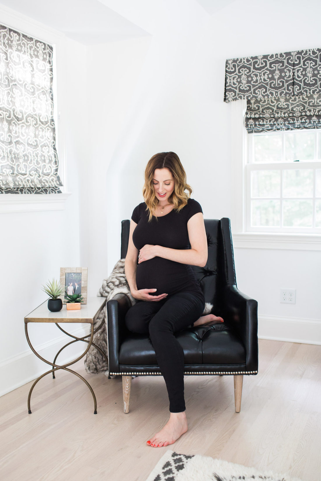 Eva Amurri Martino of lifestyle and motherhood blog REZTT cradling her baby bump in a black maternity top and jeans while sitting on a leather armchair in her master bedroom