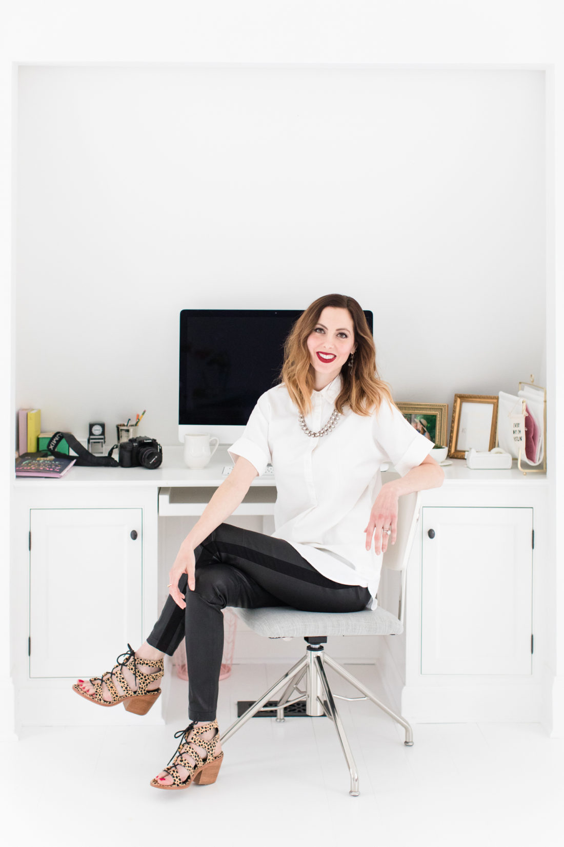 Eva Amurri Martino sits at her desk for blog REZTT, wearing a white button down shirt, leather leggings, leopard print sandals, and red lipstick
