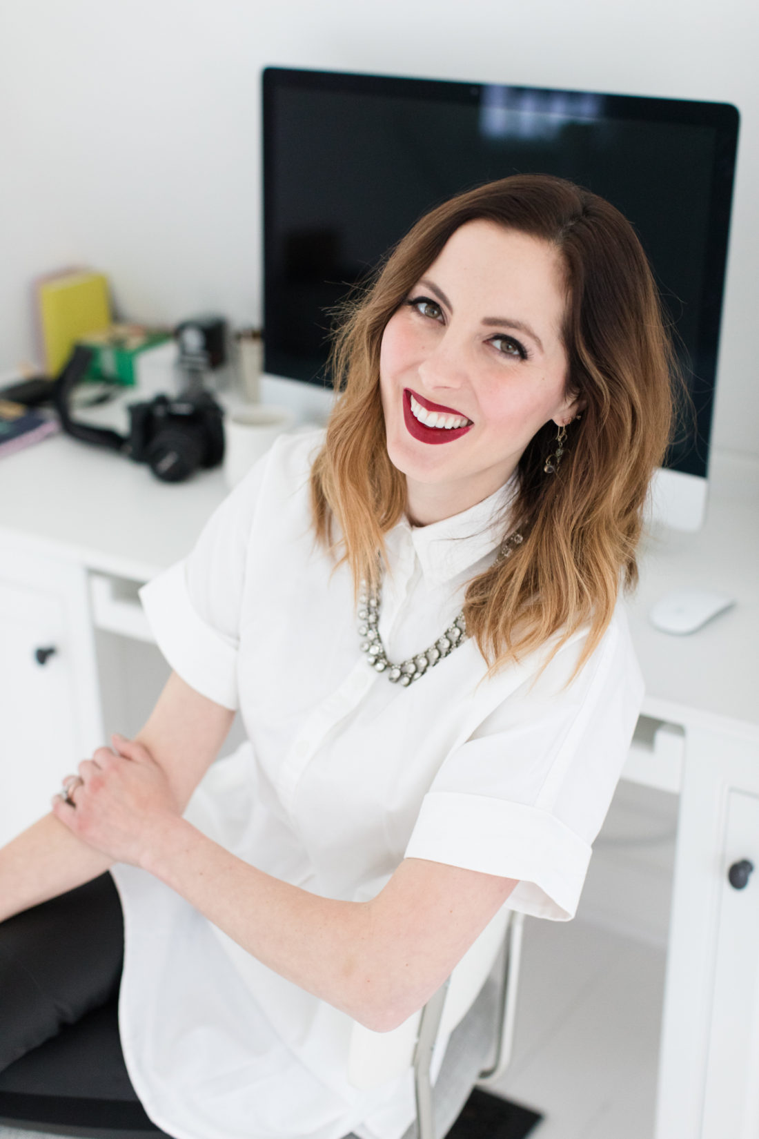Eva Amurri Martino of lifestlye and motherhood blog REZTT wears a crisp white button down shirt, a statement necklace, and red lipstick at her desk in Connecticut