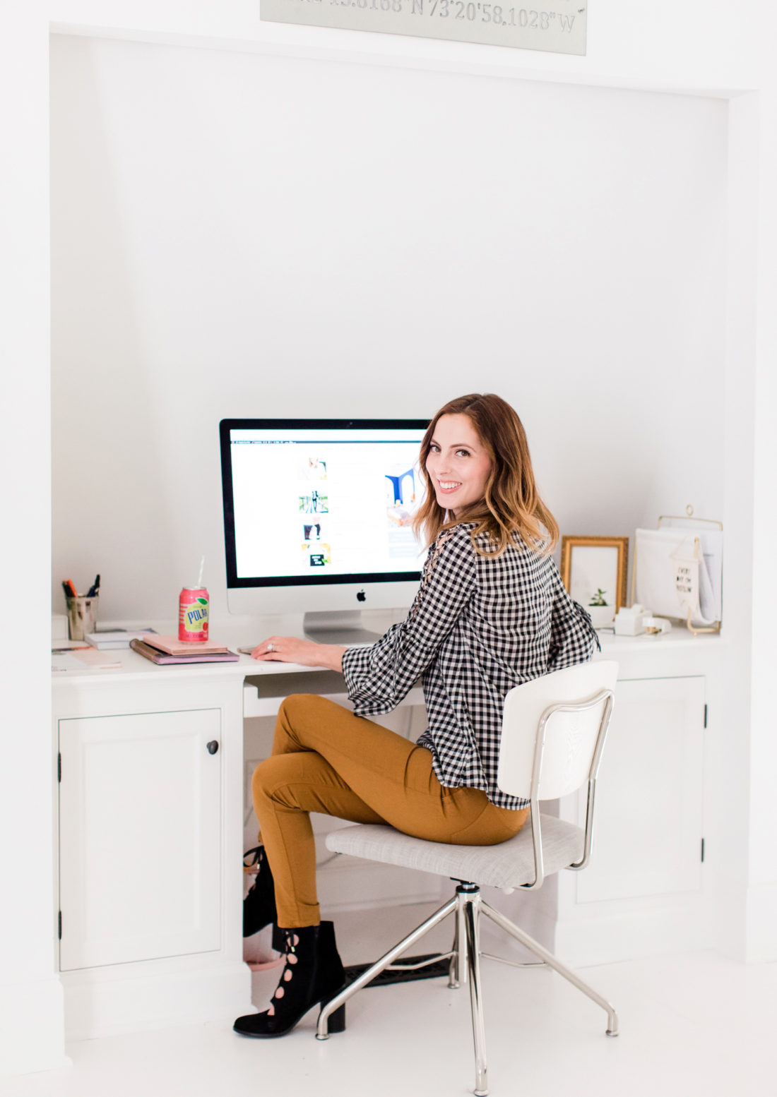Eva Amurri Martino wears tan jeans and a black and white checked top, and sits at her computer in the REZTT studio