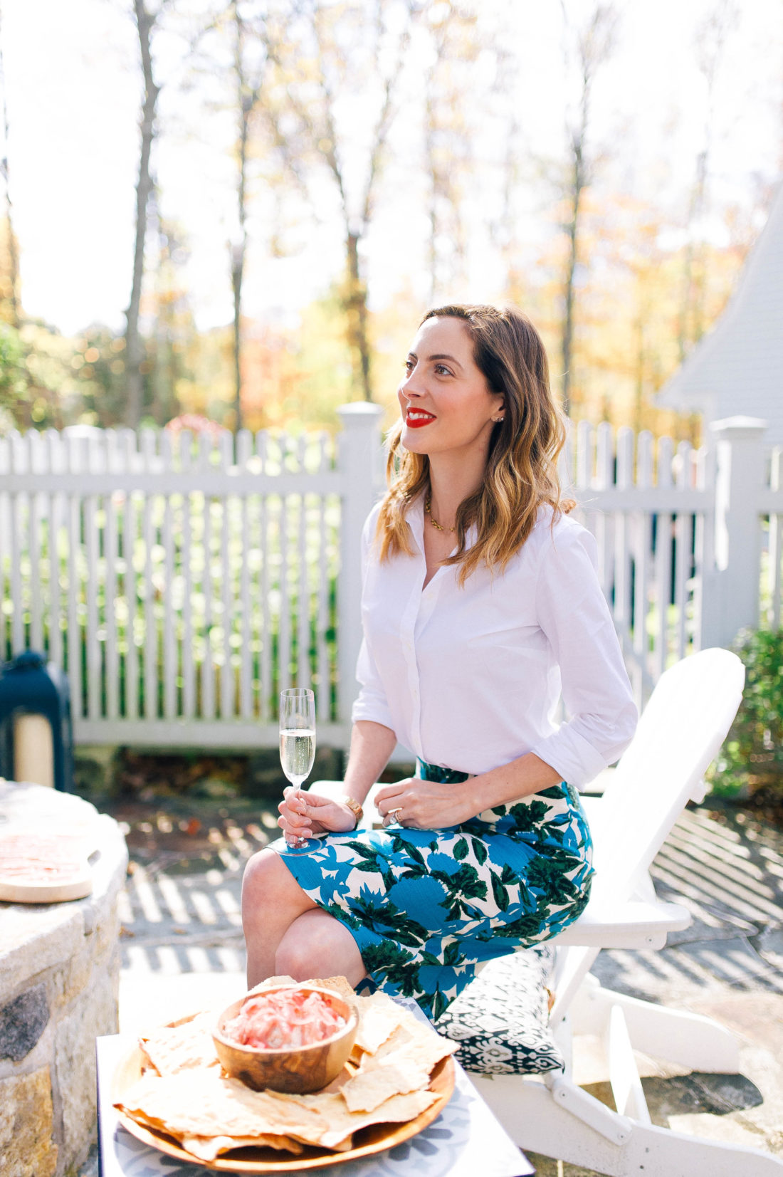 Eva Amurri Martino sits beside the fire pit at a party at her home, holding a glass of champagne and wearing a floral pencil skirt and red lipstick
