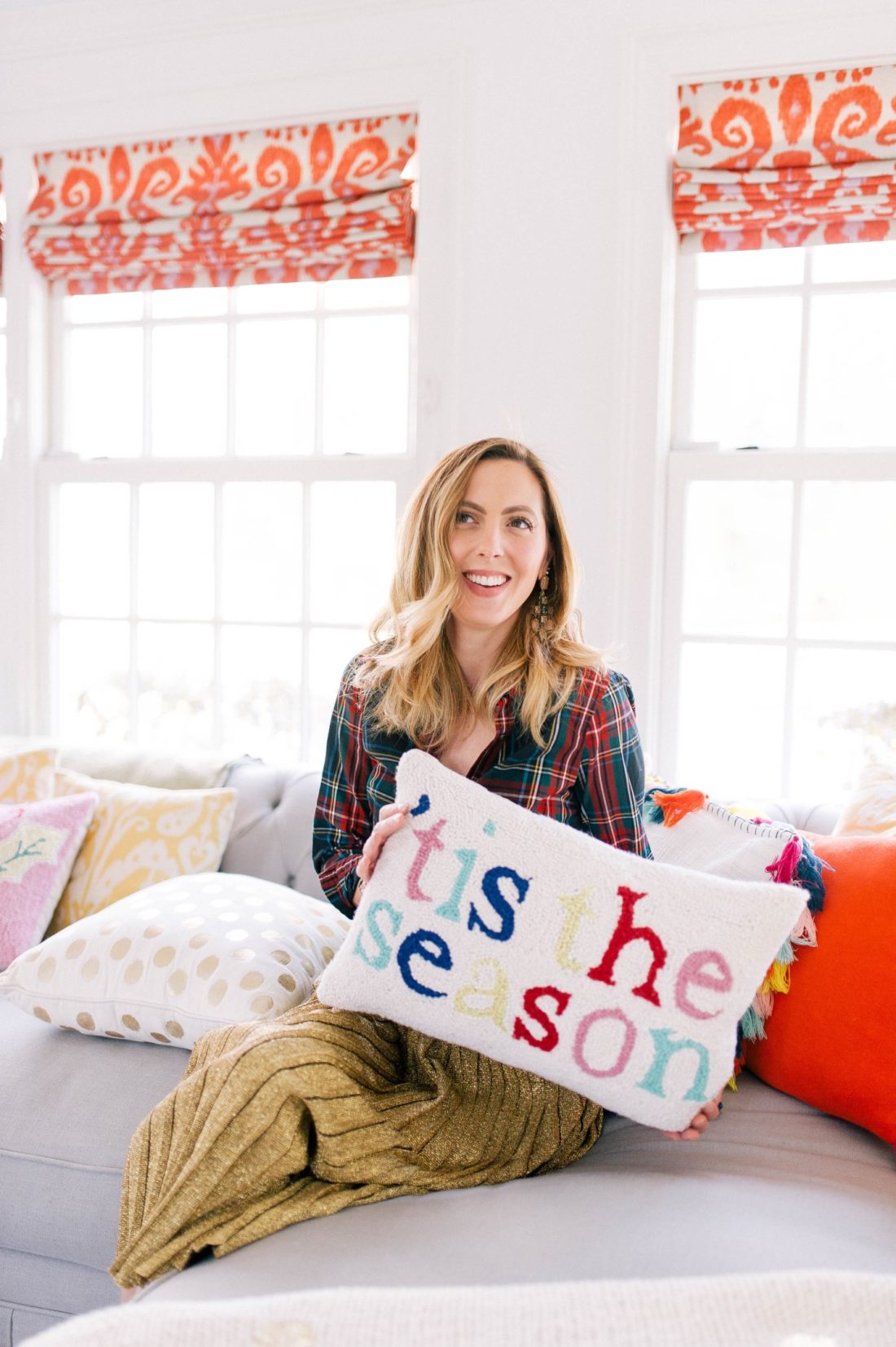 Eva Amurri Martino wears a plaid shirt and gold skirt and holds a festive Christmas pillow in the family room of her Connecticut home