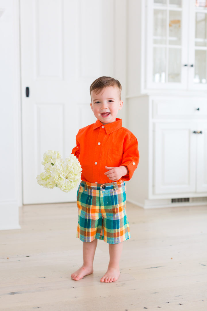 Eva Amurri Martino's son Major wears a pair of colorful green plaid shorts and an orange button up shirt while holding a handful of hydrangeas.