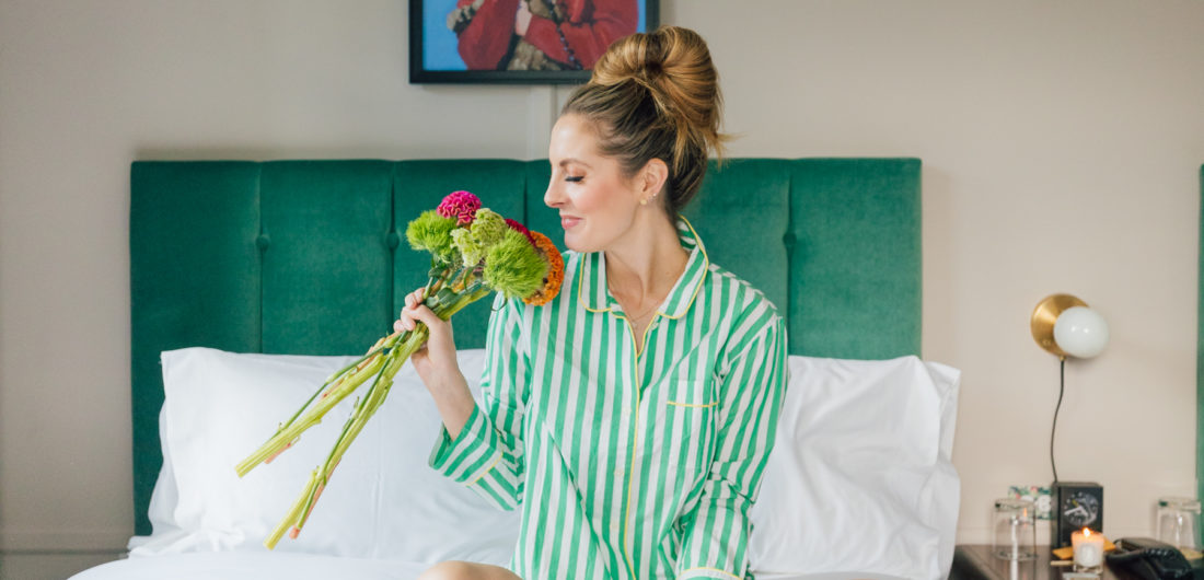 Eva Amurri Martino sits on a bed in a pair of green striped JCrew pajamas holding a bouquet of flowers